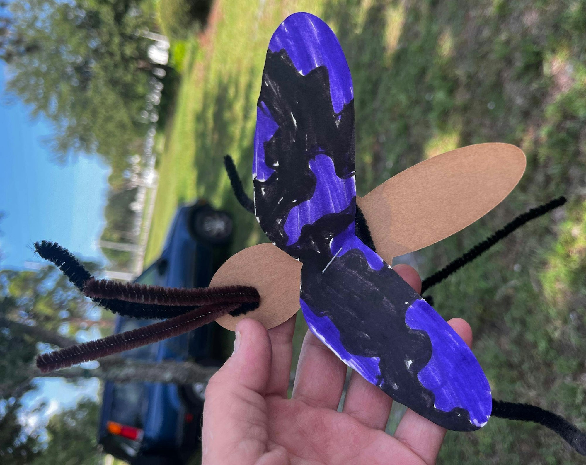 A hand holding a homemade craft butterfly with purple painted wings and black pipe cleaner antennae.