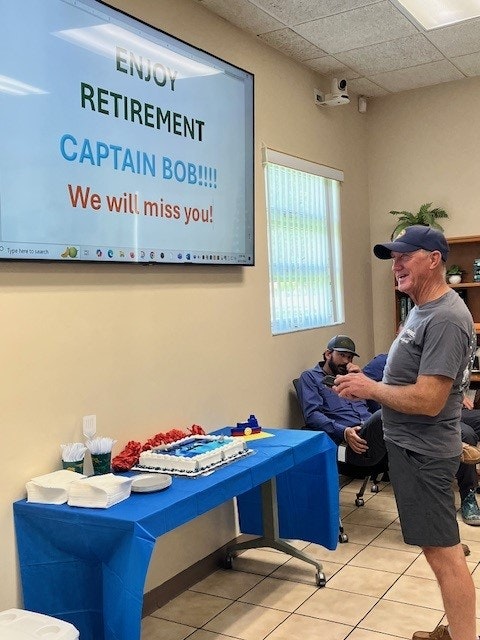 A retirement party with a smiling man, a cake, and a "Enjoy Retirement Captain Bob" presentation on screen.