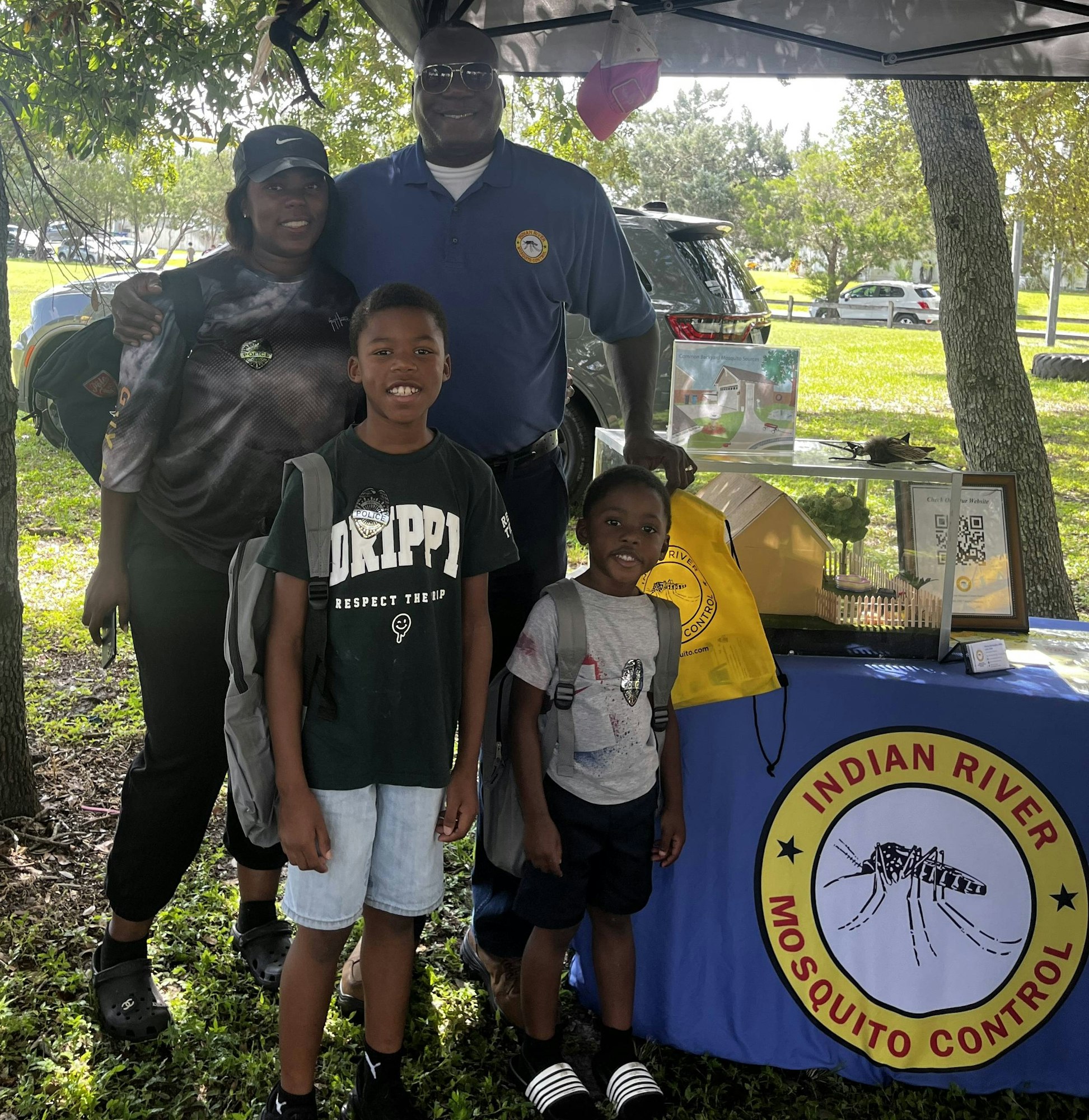 A family posing at an outdoor "Indian River Mosquito Control" event with educational materials on a table.