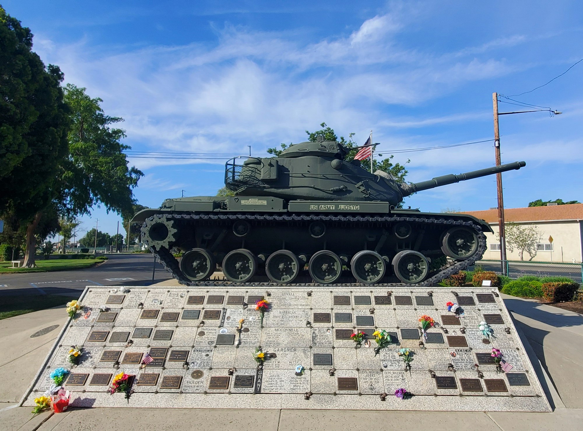 A military tank on display with a memorial wall of plaques and flowers in front.