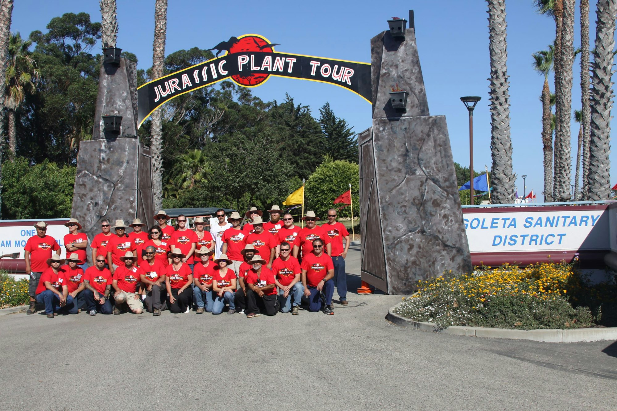 A group of people in red shirts poses in front of a "Jurassic Plant Tour" entrance sign, surrounded by palm trees.