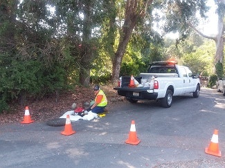 A worker in a yellow vest is on the ground near a truck, surrounded by safety cones, potentially performing maintenance.