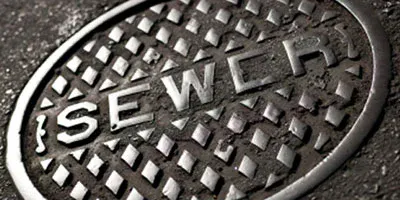 A close-up of a round, metallic sewer manhole cover with the word "SEWER" embossed on it.
