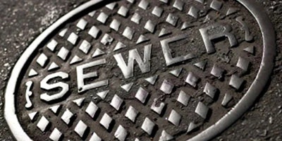 A close-up of a round, metallic sewer manhole cover with the word "SEWER" embossed on it.