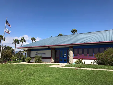 The image shows a building with a blue roof, green lawn, palm trees, and flags in front, likely a community or service center.