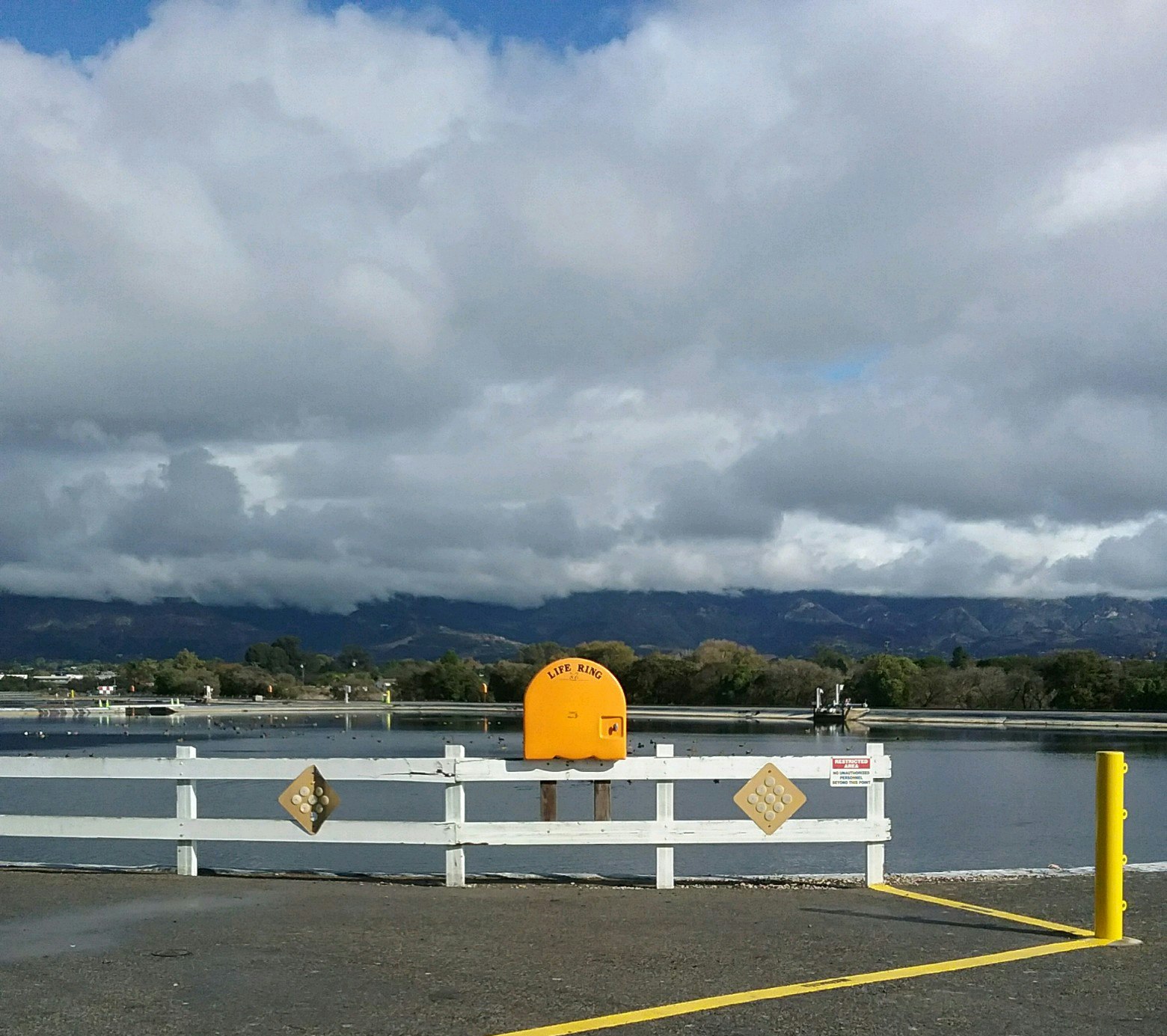 A scenic view of a body of water, mountains in the background, a white fence, and a yellow life ring sign. Cloudy sky above.