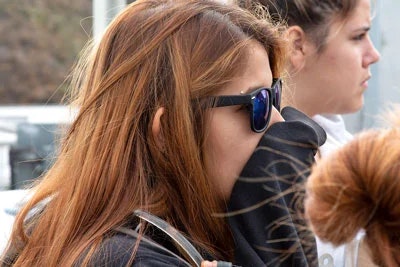 A person with long, brown hair wearing sunglasses is partially covering their face with a sleeve, while another person is in the background.