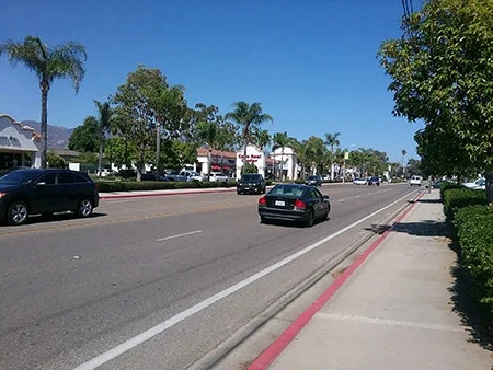A sunny street scene with cars, palm trees, and shops lining the road, showcasing a vibrant suburban atmosphere.