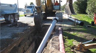 A construction site with heavy machinery digging and laying a large pipe in a trench along a roadway.