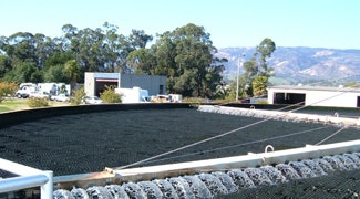 The image shows a large circular water treatment facility with black filtration media and surrounding trees and buildings.