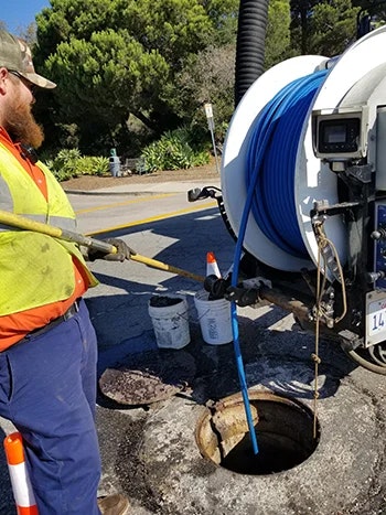 A worker in a safety vest and cap is using equipment to inspect or clean a sewer line, with a hose and bucket nearby.