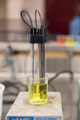 The image shows a beaker with yellow liquid and two electrodes for measuring conductivity. It’s set on a lab stirrer.