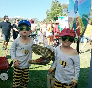 Two kids in matching outfits and sunglasses pose with a toy dinosaur at a lively outdoor event filled with people.