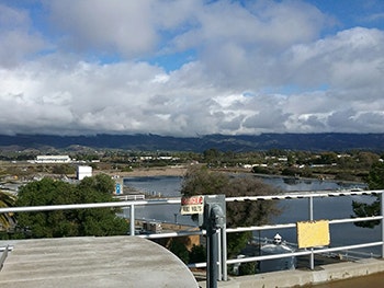 A scenic view of a river flanked by low hills and buildings, with a partly cloudy sky above.