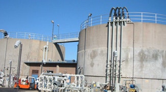 The image shows industrial storage tanks with pipes and equipment, set against a clear blue sky.