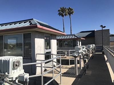 The image shows a modern building with a metal roof, surrounded by equipment and palm trees under a clear blue sky.