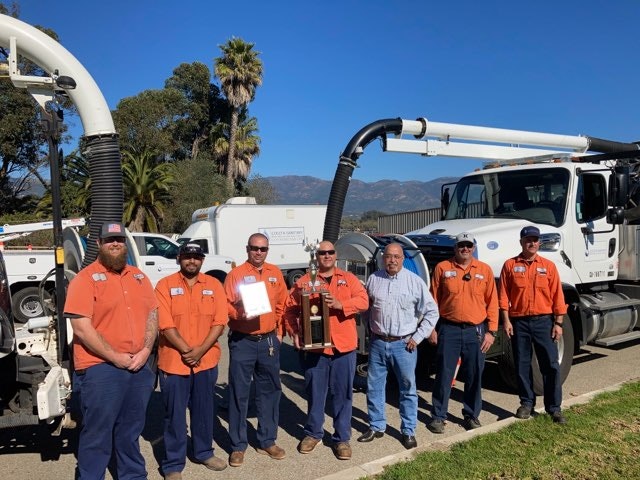 A group of workers in orange uniforms stands with a trophy and certificate in front of service trucks in a sunny setting.