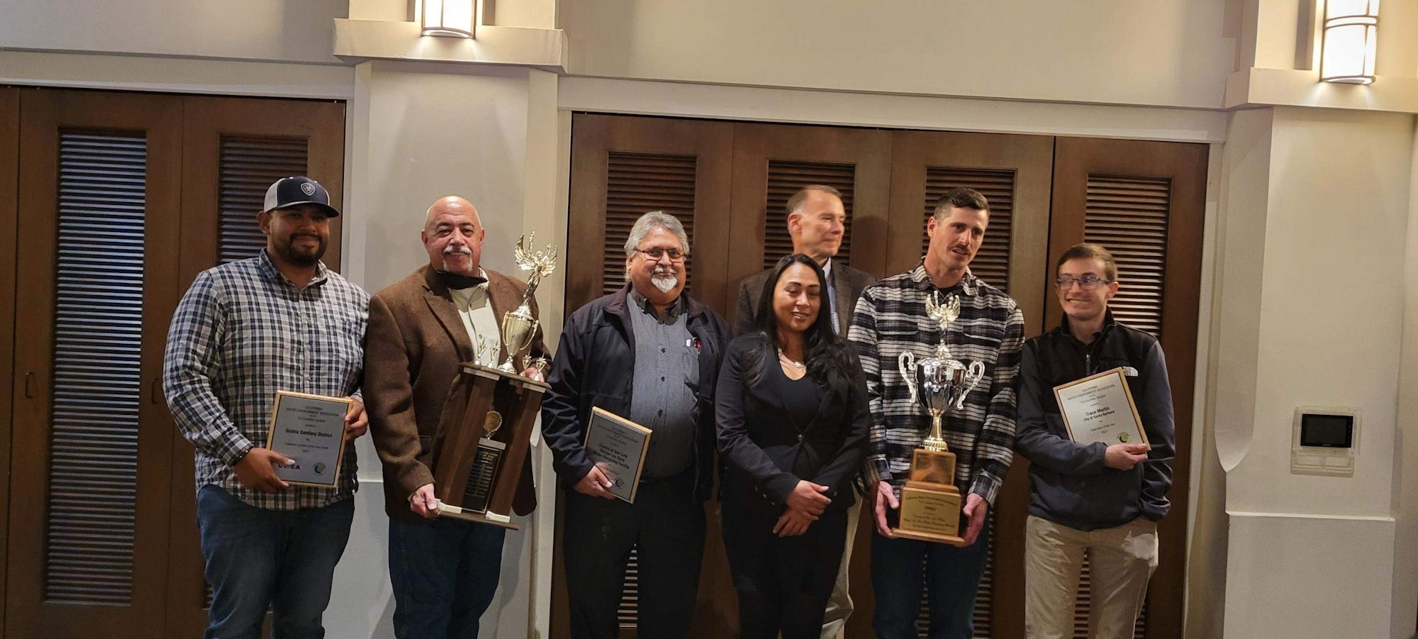 A group of seven individuals poses together, holding trophies and awards, likely celebrating an achievement or competition.