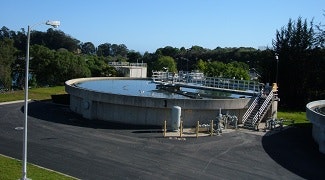 The image shows a wastewater treatment facility with large circular tanks and surrounding greenery under a clear blue sky.