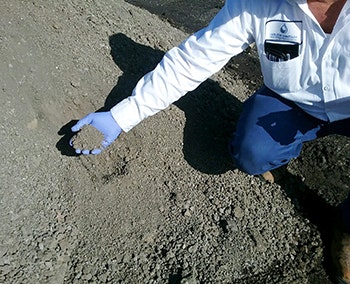A person in a blue glove is kneeling and holding a handful of gravel or soil, inspecting it closely.