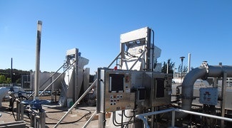 The image shows industrial equipment, possibly part of a processing facility, under a clear blue sky with some pipes and control panels.