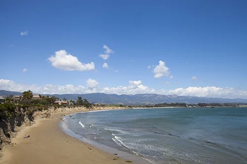 A scenic beach view with gentle waves, sandy shore, distant mountains, and a clear blue sky dotted with fluffy clouds.