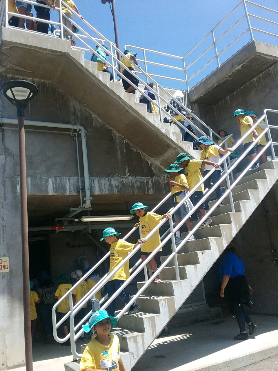 A group of children in yellow shirts and blue hats is descending a staircase at a building or facility, enjoying an outing.