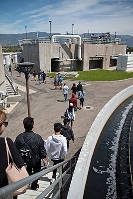 People walking towards a large industrial facility, likely a water treatment plant, with tanks and pipes visible.