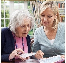 A younger woman is assisting an older woman with calculations using a calculator at a table in a bright, homey setting.