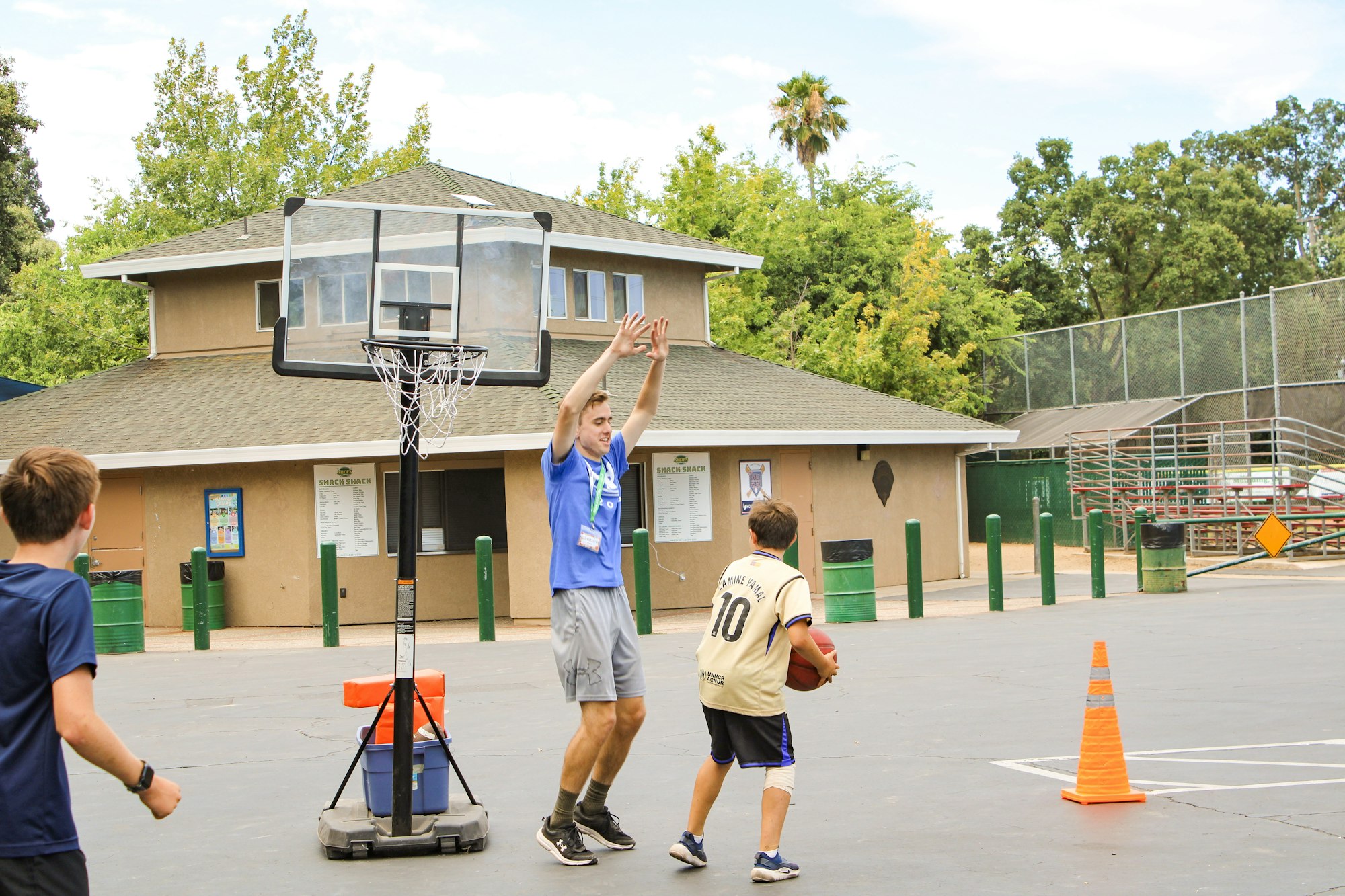 A basketball game in progress with a coach and kids on a court, featuring a hoop, playground, and nearby buildings.