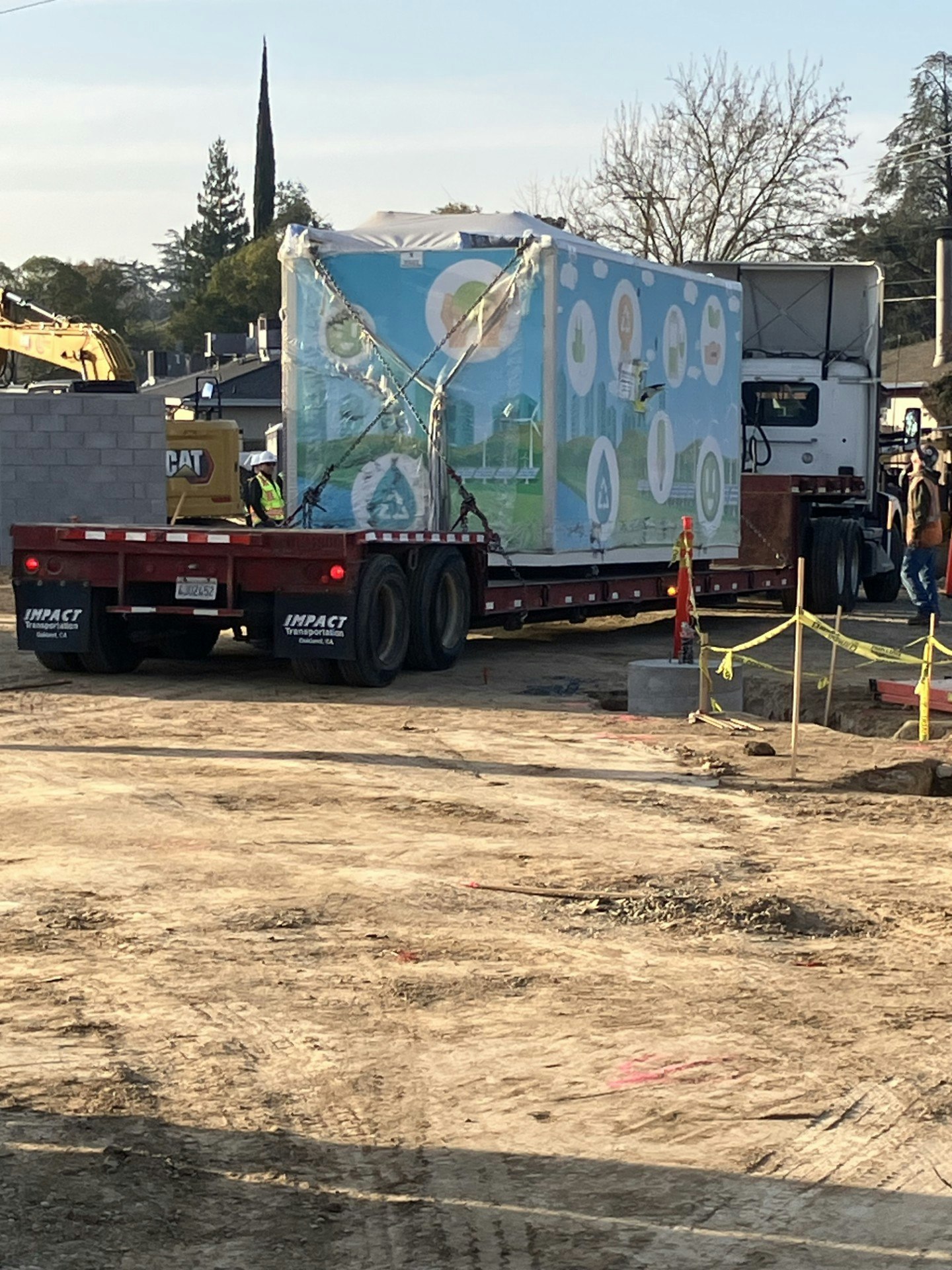 A truck is unloading a large, brightly decorated container at a construction site, with workers and machinery nearby.
