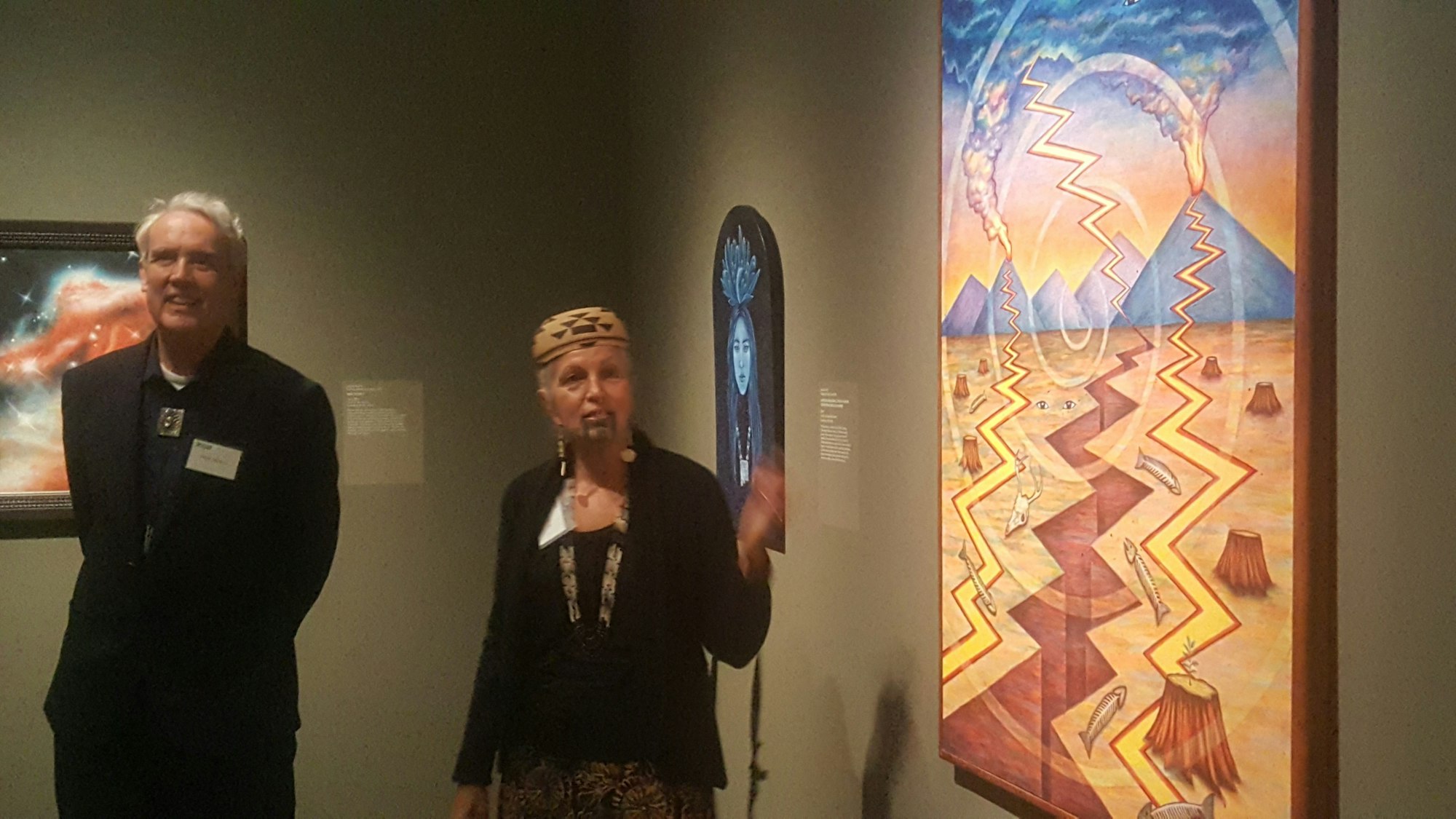 The image shows two people in an art gallery, one speaking, with colorful abstract artwork in the background.