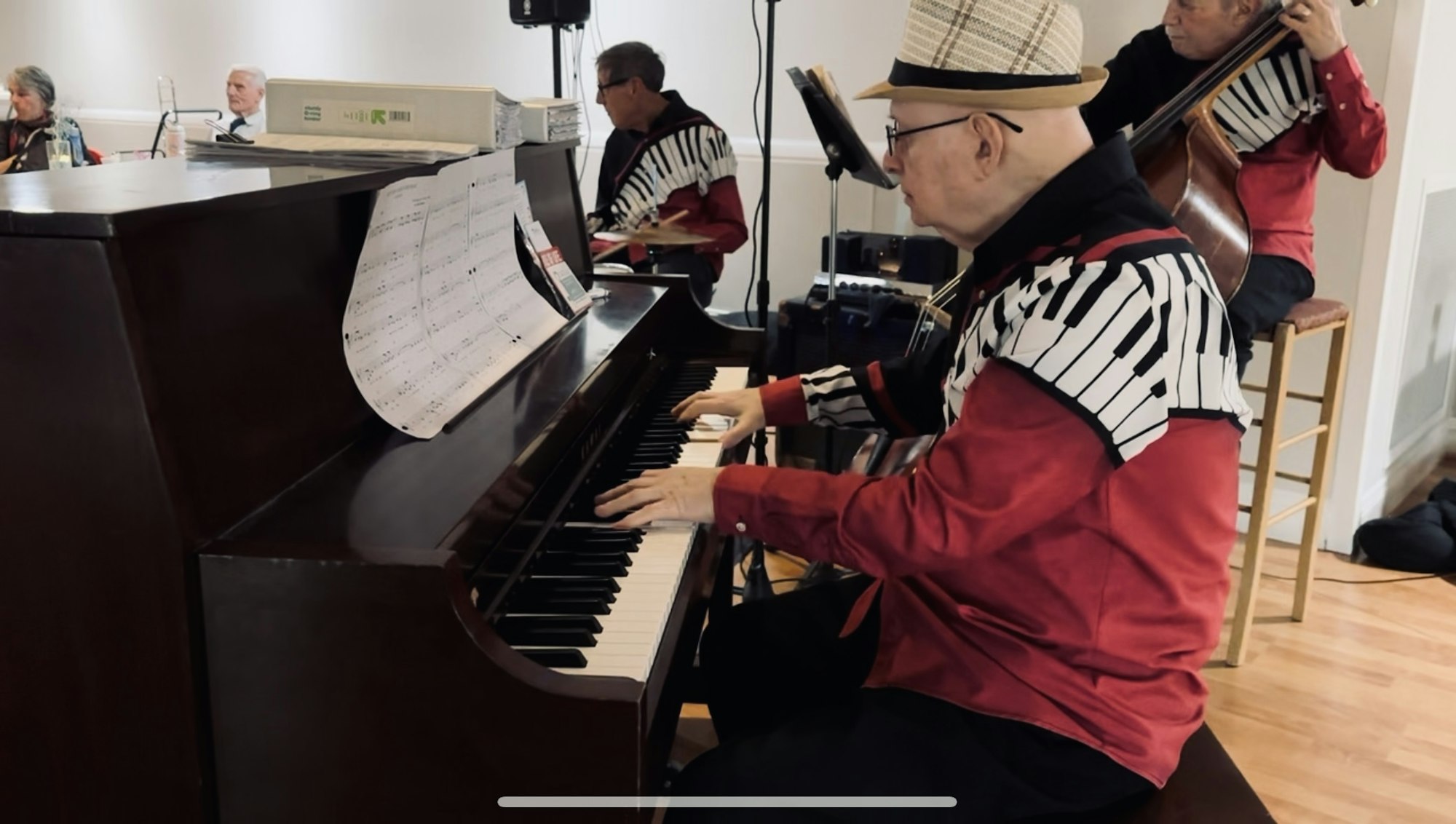 A musician plays the piano, wearing a shirt with piano key patterns. Two other musicians are in the background.