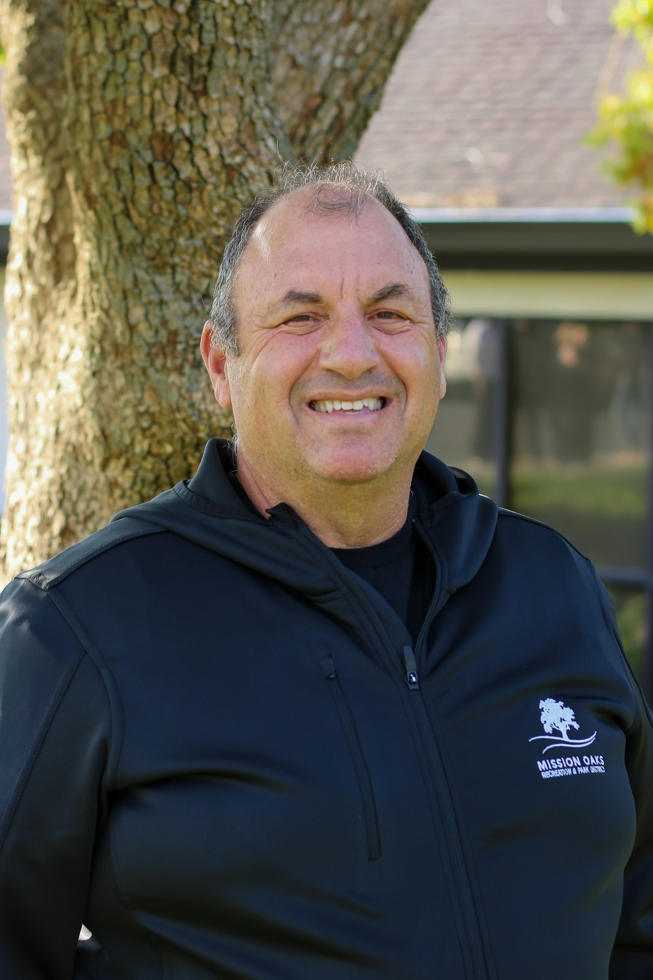 A person smiles outdoors, wearing a black jacket with "Mission Oaks Recreation & Park District" logo, standing in front of a tree.