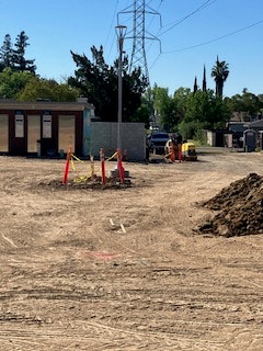 An empty lot with dirt and debris, construction barriers, a few workers, and nearby trees and power lines in the background.