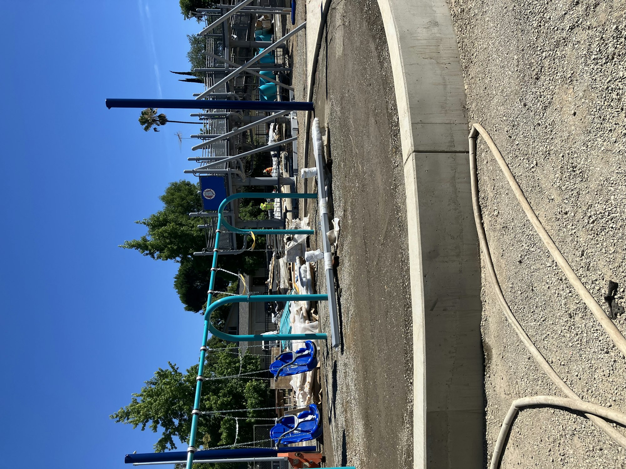 The image shows a playground under construction with blue playground equipment and a clear sky.