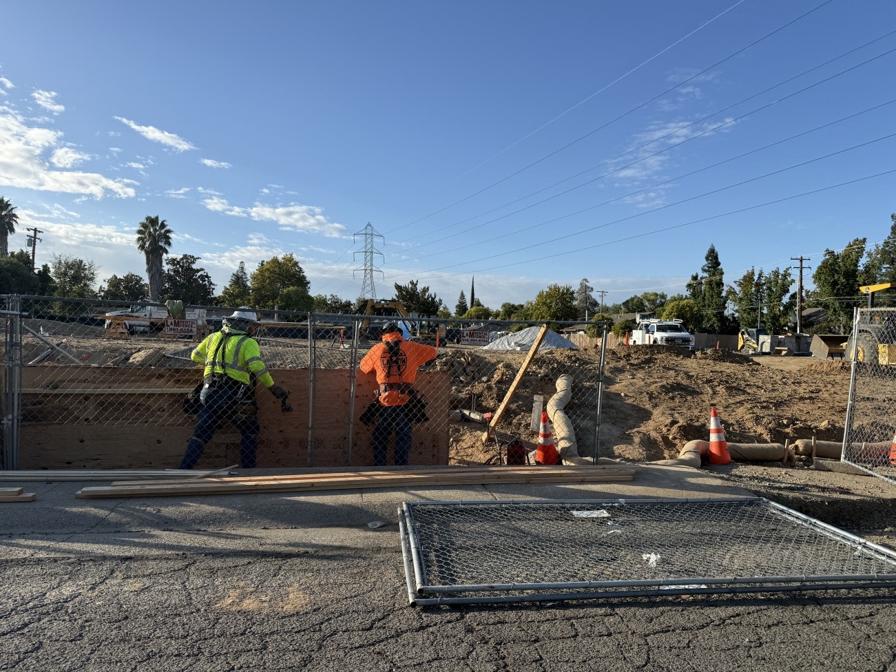 emPOWERment Park construction site with two workers in a fenced-off area, piles of dirt, and construction vehicles in the background.