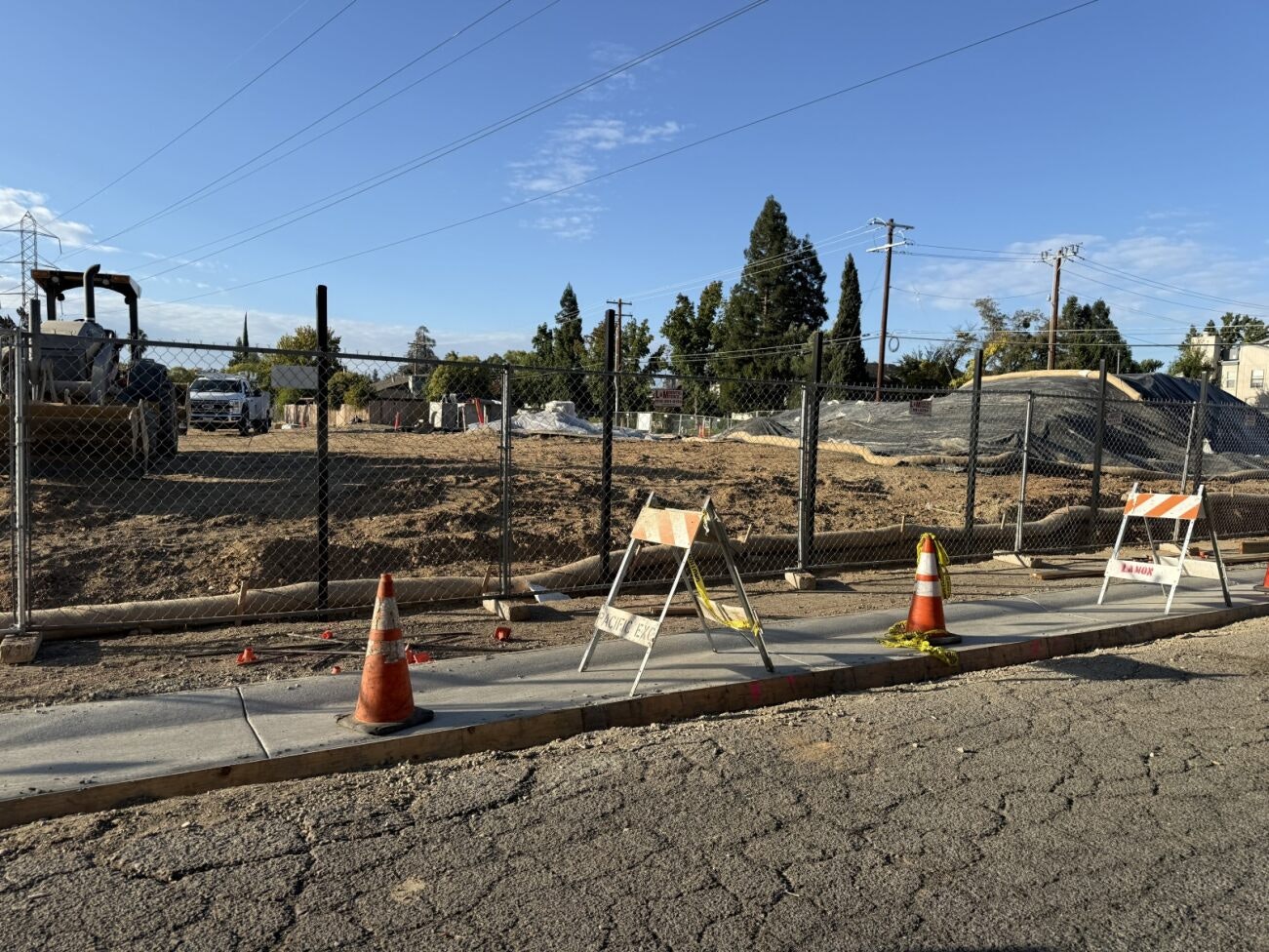 emPOWERment Park Construction site with tractors, machinery, fenced area, traffic cones, and barricades on a cracked road under a clear sky.