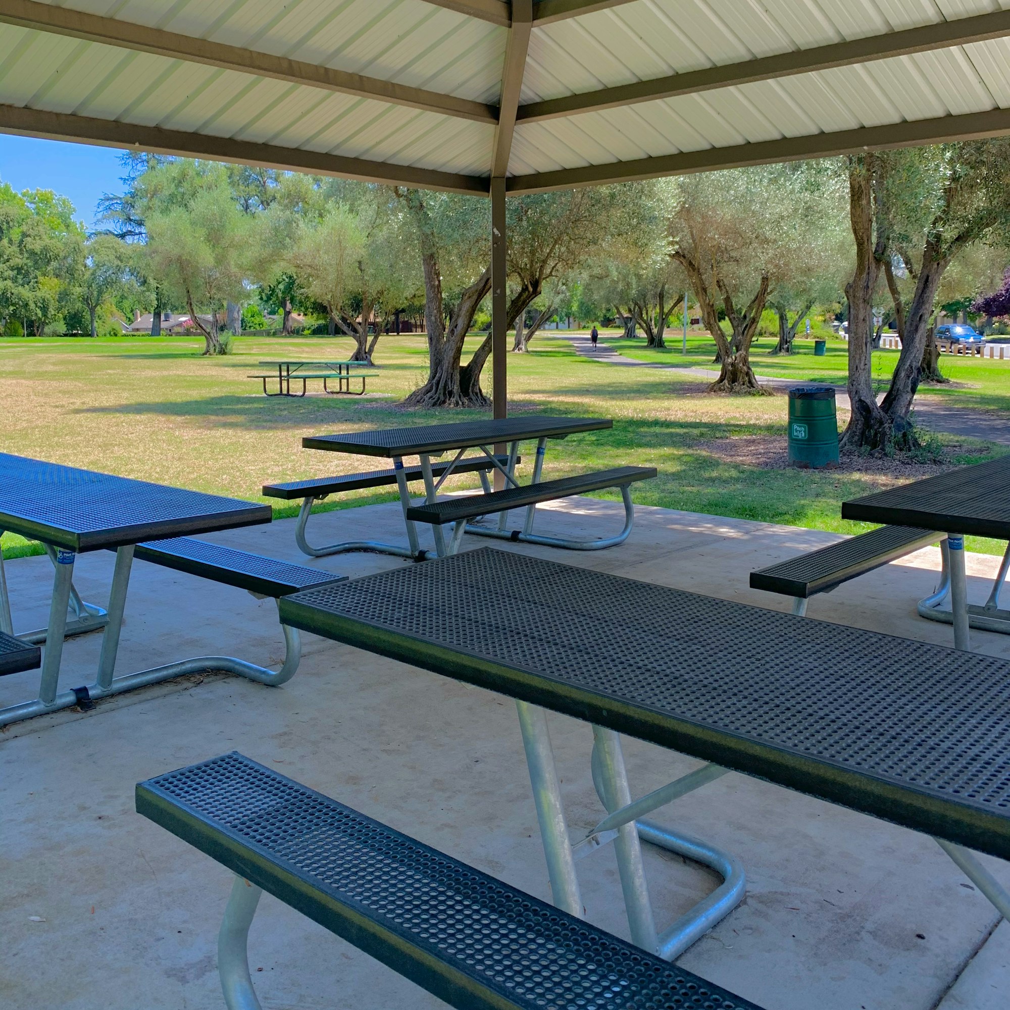 A covered picnic area with metal tables and benches, surrounded by grassy fields and trees in a sunny park setting.