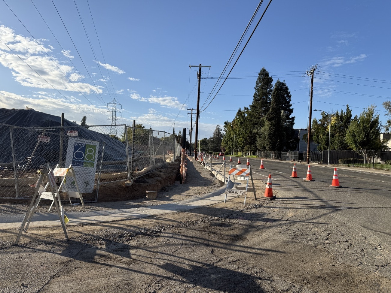 The Bell Way sidewalk along side emPOWERment Park under construction with traffic cones and barriers alongside a fenced-off area, under a clear blue sky.