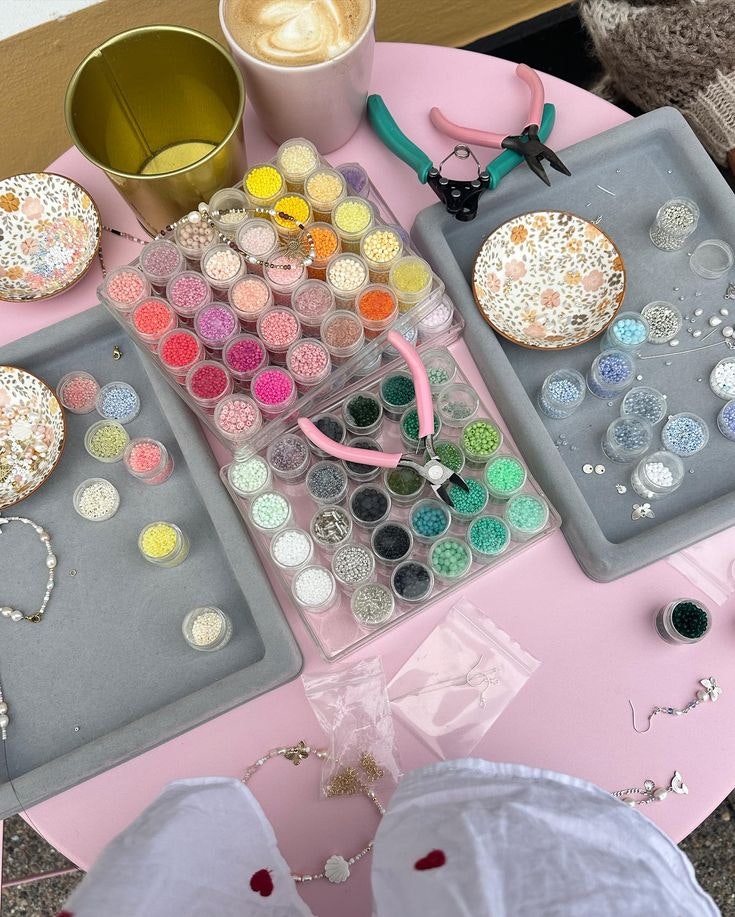 Jewelry-making setup with colorful beads, tools, a latte, and decorative plates on a pink table.