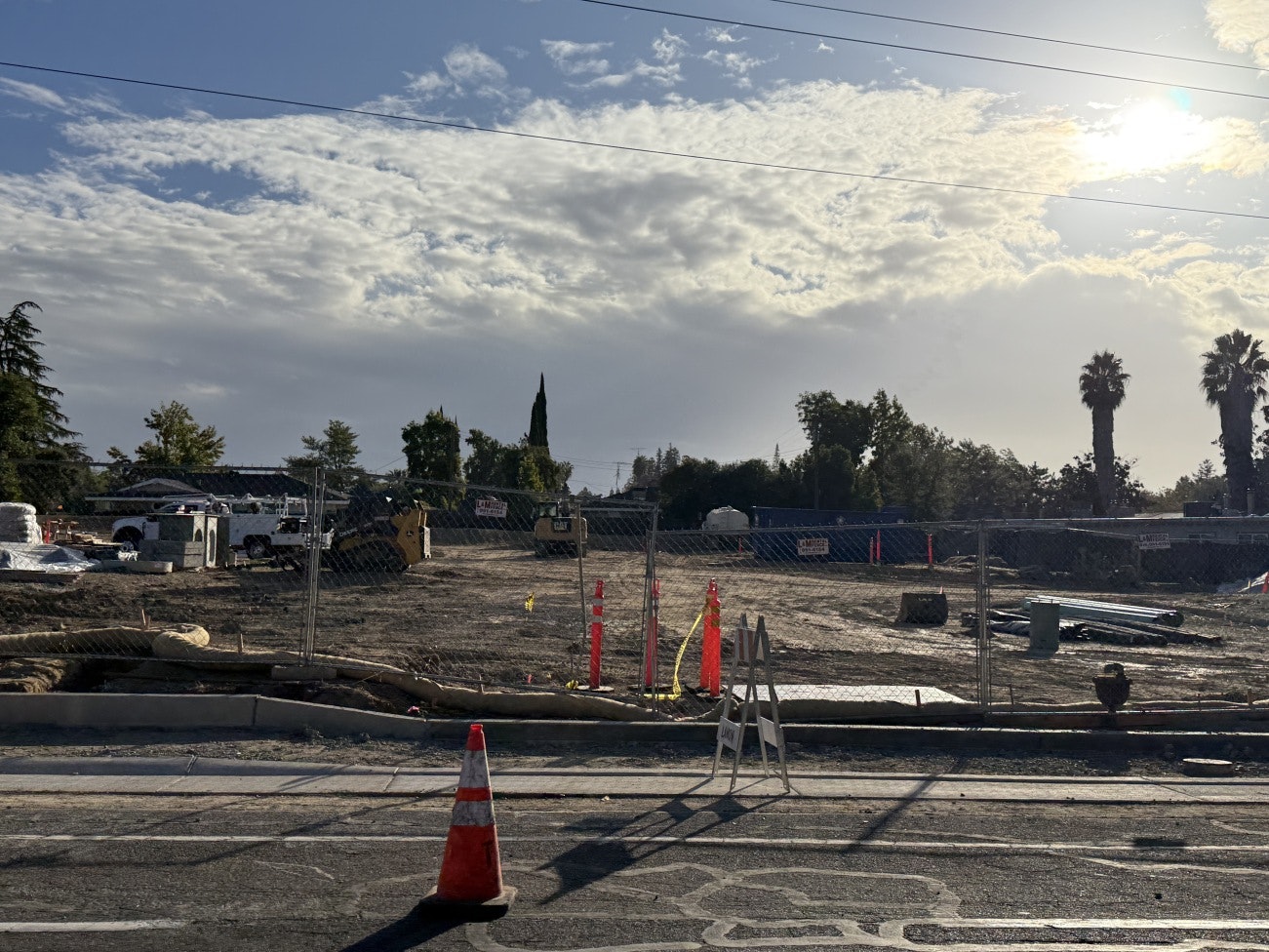 emPOWERment Park Construction site with vehicles, fencing, and traffic cones under a cloudy sky with the sun.