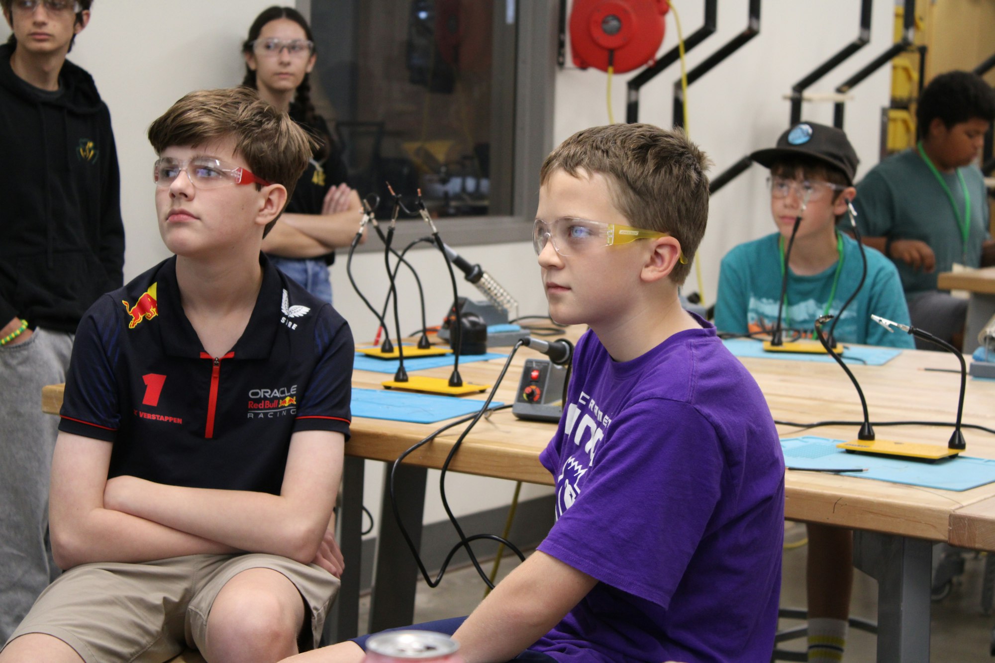 Students wearing safety goggles listen attentively in a classroom with workstations for projects, showing engagement in hands-on learning.