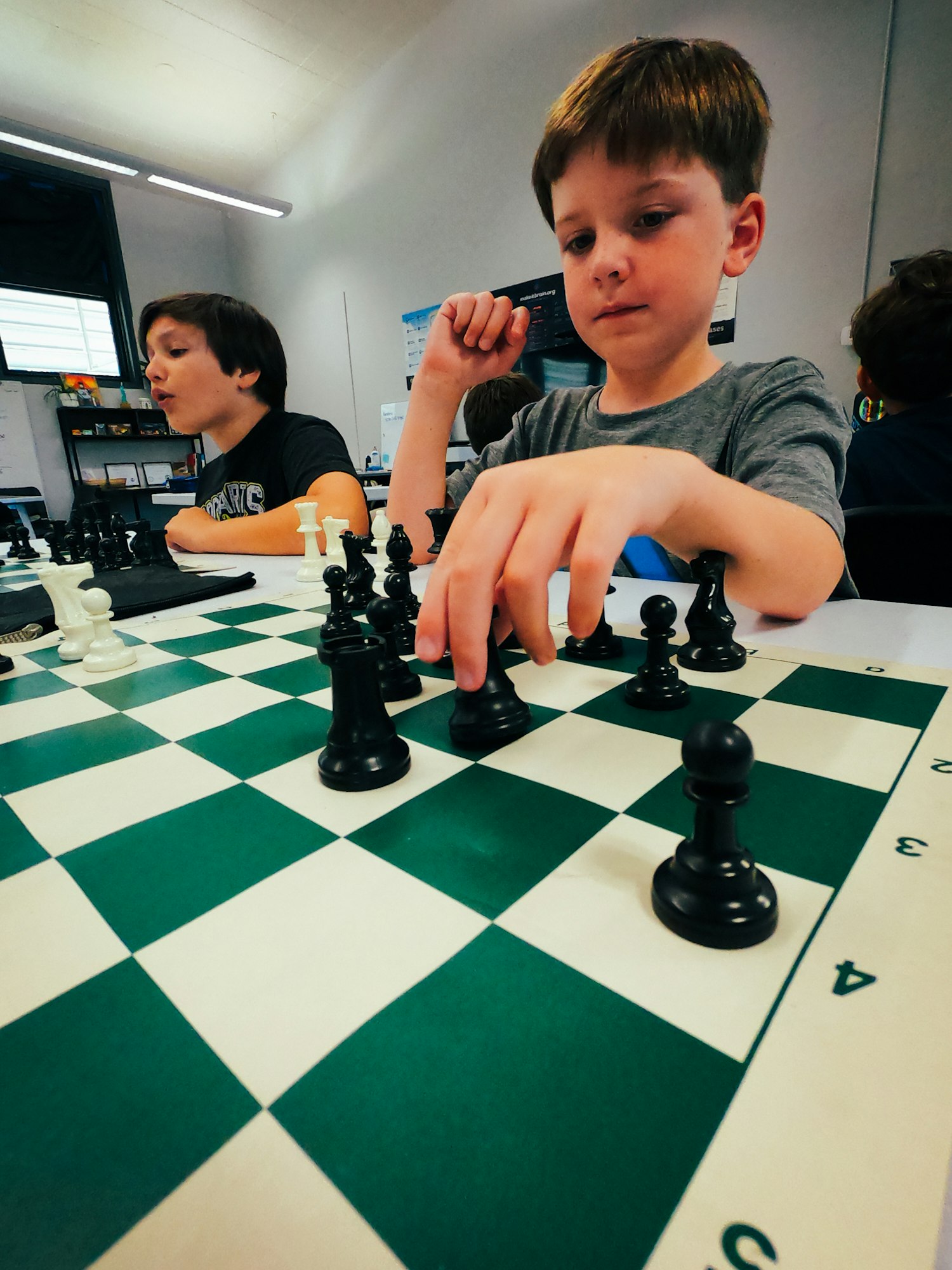 Two kids playing chess, one focusing on moving a piece, the other watches intently.