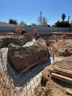 The image shows a construction site with workers, large rocks, and reinforcement materials for a concrete foundation.