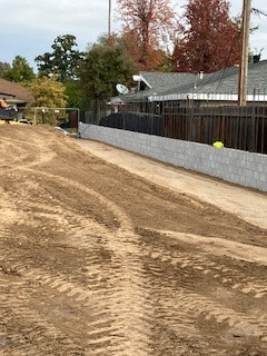 Dirt lot with tire tracks, concrete wall, houses, and trees in the background.