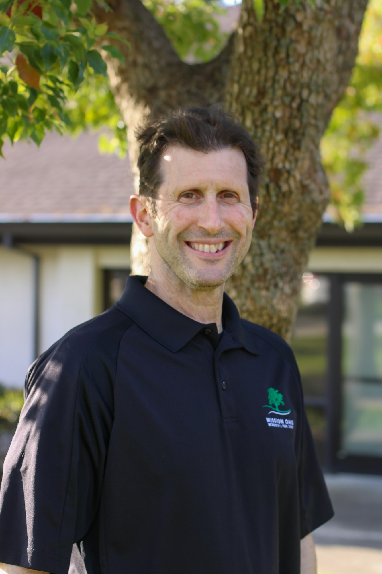 A person smiling outdoors in front of a tree, wearing a black polo shirt.