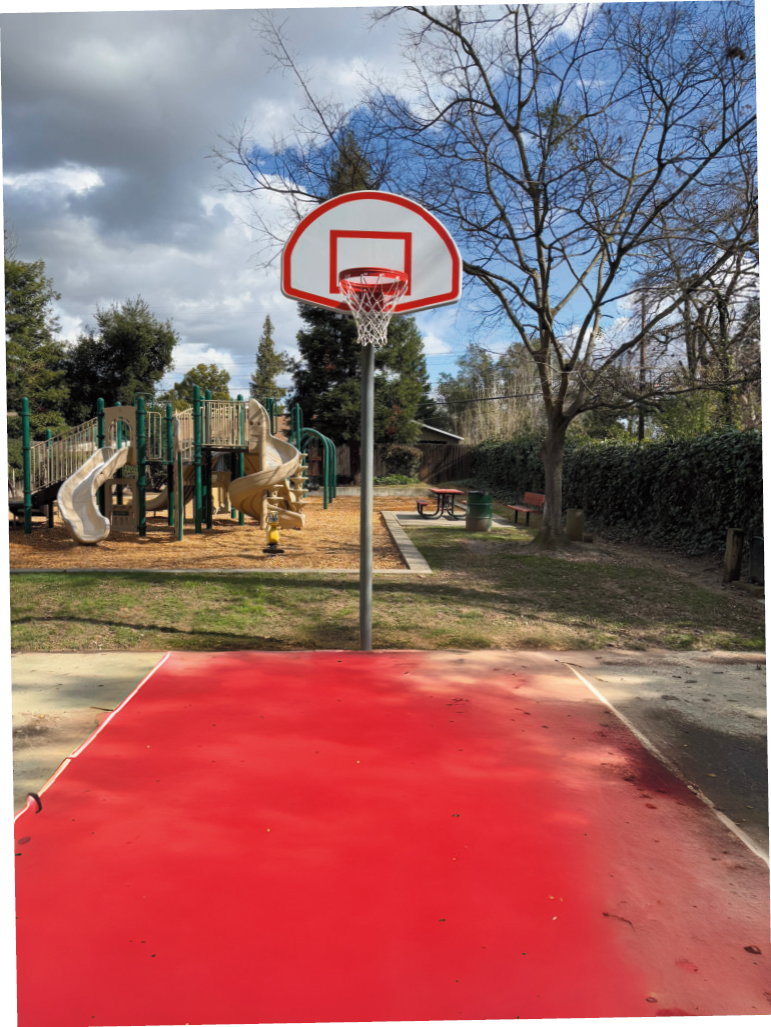 Basketball hoop on a red court with a playground and trees in the background under a cloudy sky.