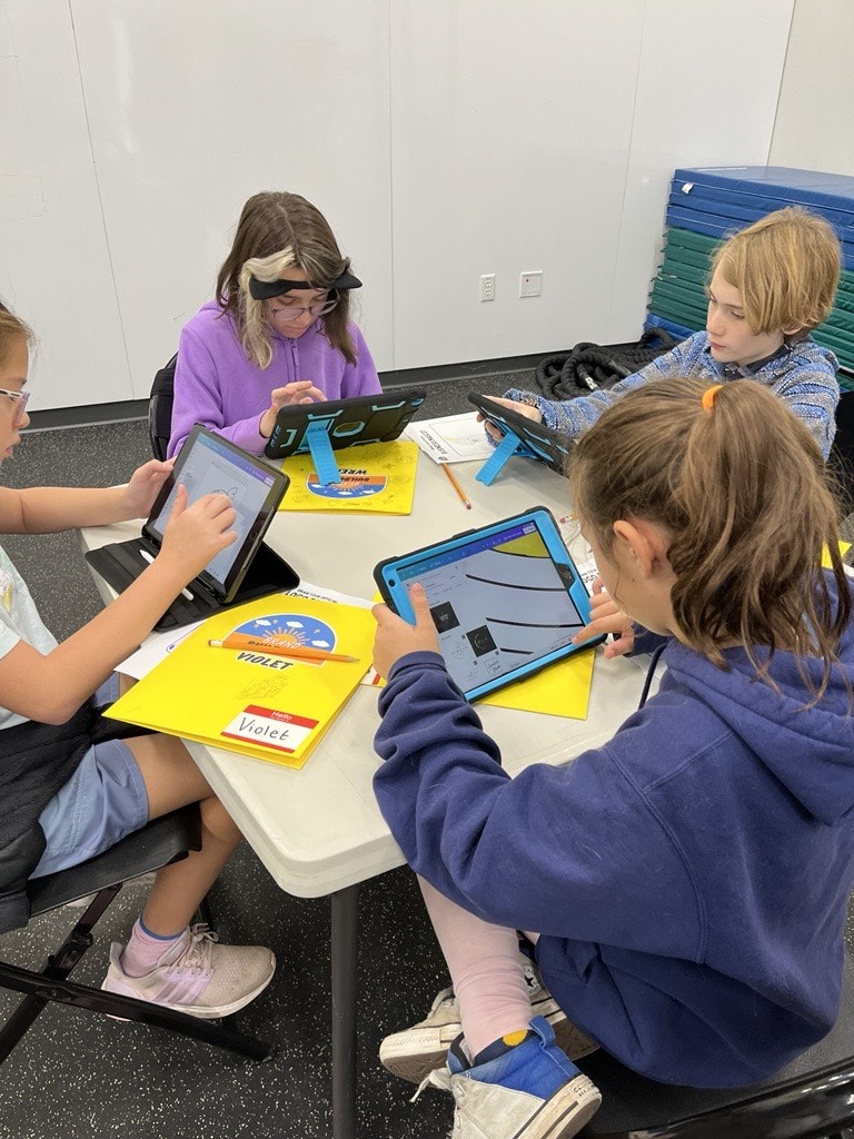 Four children sitting at a table, each using a tablet while working on assignments or activities. Colorful folders are visible.