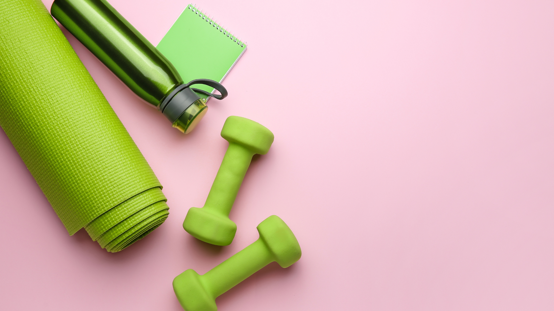 Green yoga mat, water bottle, notebook, and dumbbells on a pink background.